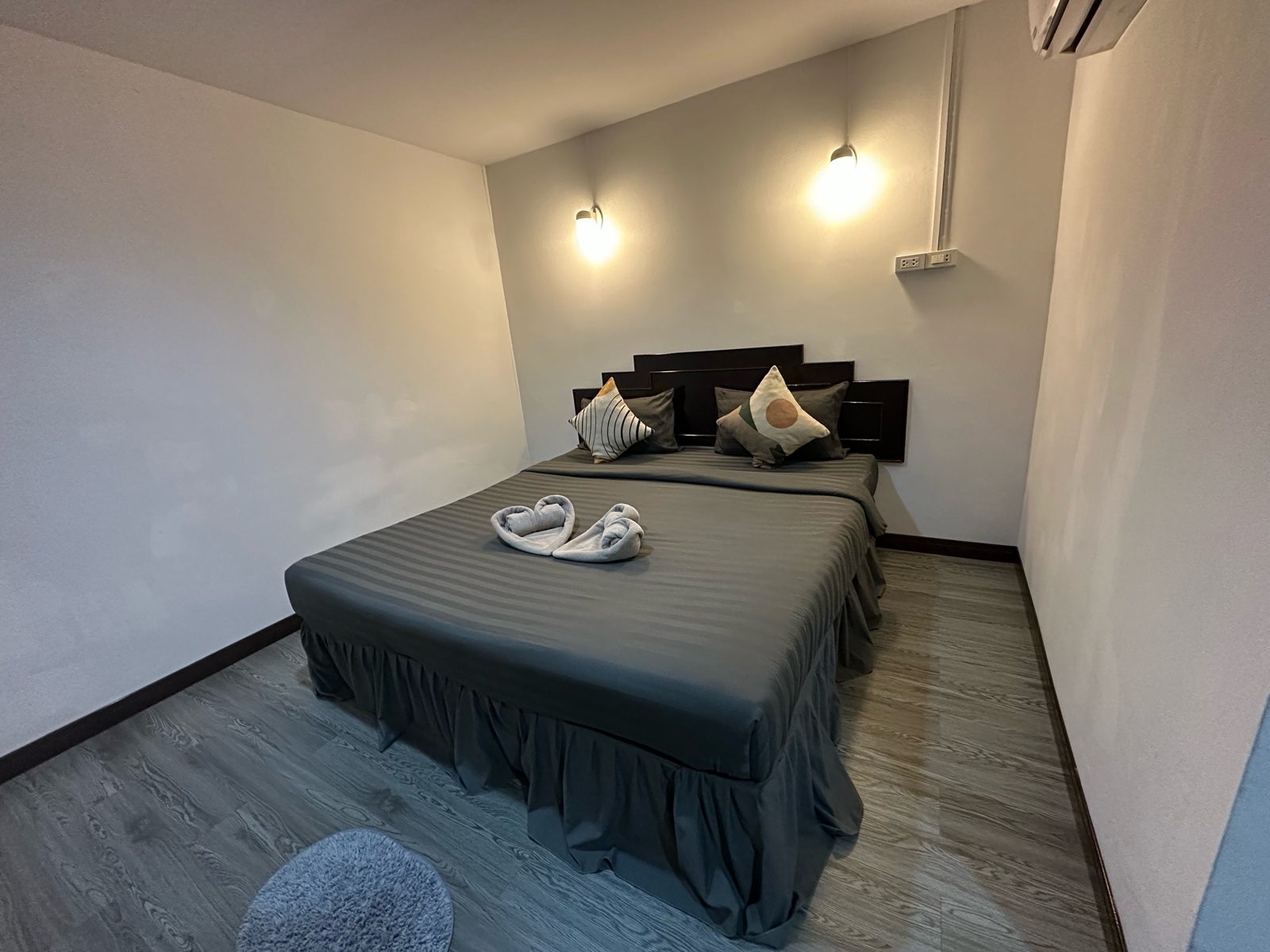 Bedroom with a grey striped bed, dark wooden headboard and two wall lamps, straight off the phone. The far wall and the floor behind the bed sit in shadow.